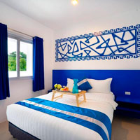 A neatly made bed with blue and white bedding, a blue headboard, and a tray with coffee service on it. The room has a window with curtains and a decorative wall panel above the bed.