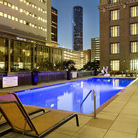 Outdoor pool on a high-rise building's terrace surrounded by city lights and buildings at dusk. Two brown lounge chairs are in the foreground beside the pool.