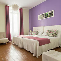 A bedroom with two single beds, white and pink bedding, a purple accent wall, pink curtains, a window, a floral artwork, and brown wooden flooring.