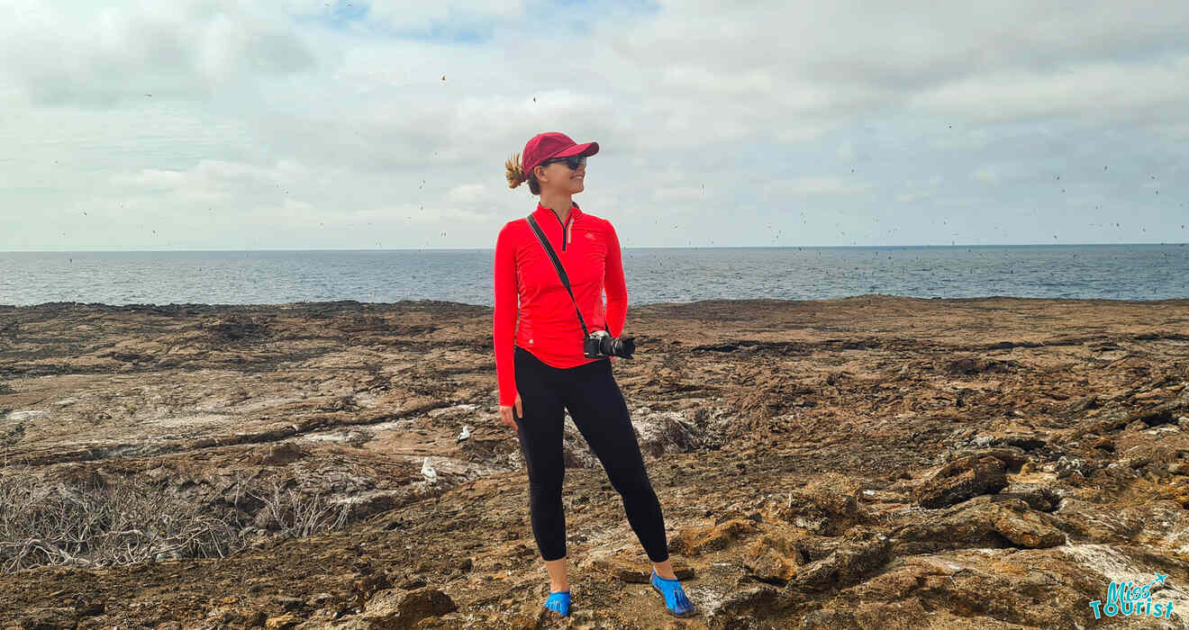 author of the post in a red long-sleeve shirt, black pants, and blue shoes, wearing a red cap and sunglasses, stands on a rocky coastal area with a camera hanging by their side. The ocean is visible in the background.