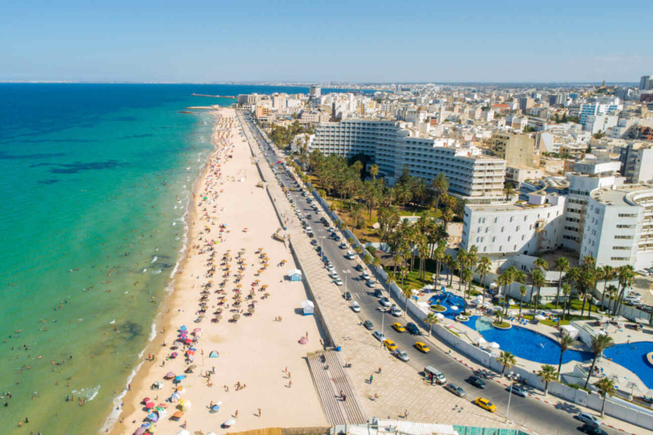 Aerial view of a bustling Tunis beach with turquoise waters, umbrellas dotting the sandy shore, and a busy street lined with hotels and buildings