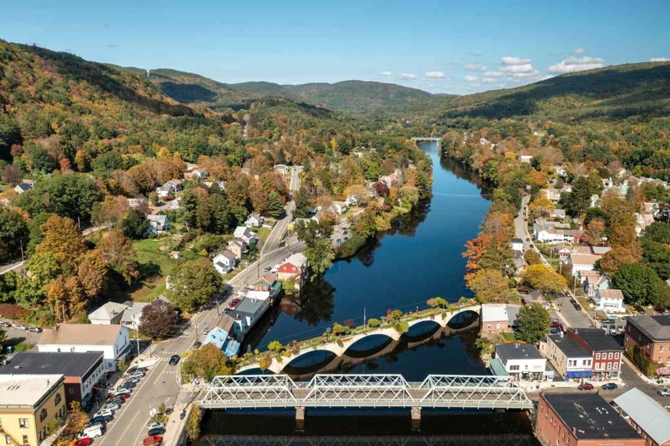 Aerial view of a small town with a river running through it, surrounded by trees with autumn foliage, several buildings, and bridges crossing the river.