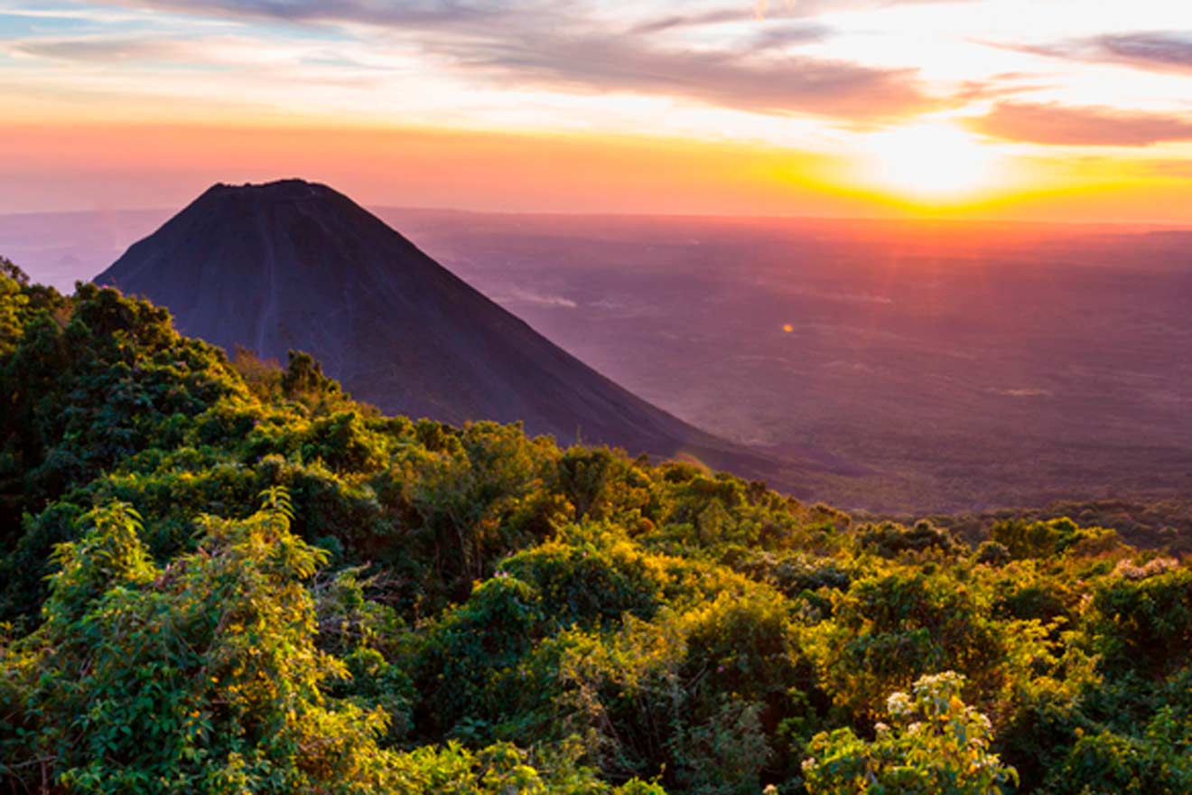 Sun setting behind a prominent conical volcano with lush green vegetation in the foreground. The sky is vibrant with shades of orange, pink, and purple.