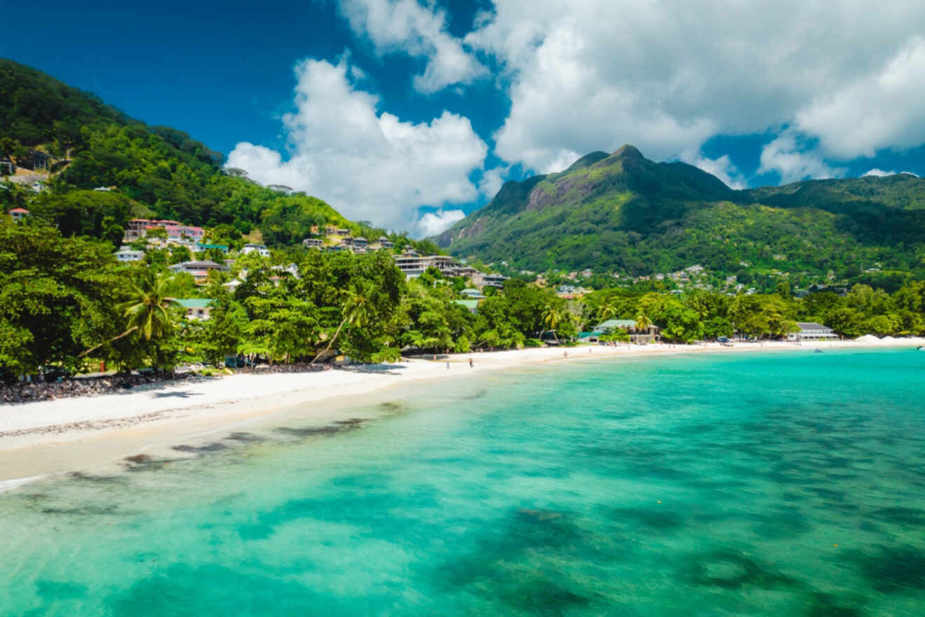 A pristine beach with turquoise water, backed by lush green hills and small buildings nestled among the trees, under a bright blue sky with fluffy clouds.