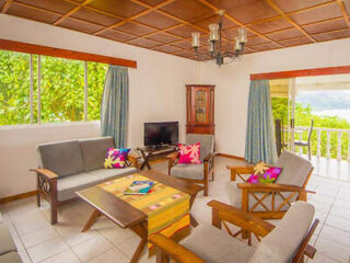 A bright living room with wooden furniture, a cushioned sofa set, colorful pillows, and a TV, with large windows and a door leading to a balcony.