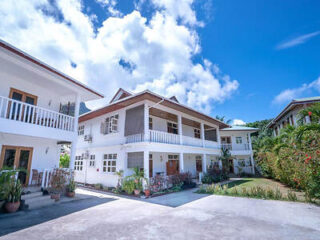 A two-story white building with balconies, set against a bright blue sky, surrounded by tropical greenery.