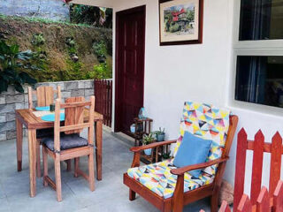 A cozy outdoor patio with a small wooden table and chairs, along with a colorful cushioned chair, bordered by a red picket fence.