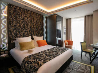 A modern hotel room with a double bed, patterned headboard, two pillows, an orange cushion, a small desk, a chair, and a window with curtains.