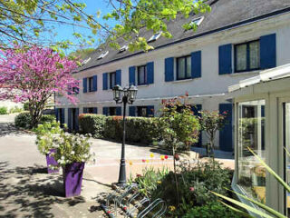 A white two-story building with blue shutters is surrounded by greenery and colorful plants. A black lamppost stands near a bike rack in the foreground. Pink blossoms are visible on the left.