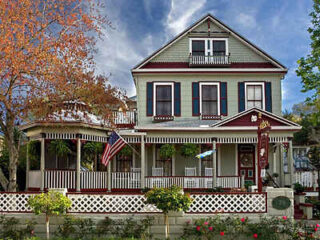 Victorian-style house with a wraparound porch, hanging plants, and American flag. The house is green with red trim and white accents, surrounded by trees and a picket fence.