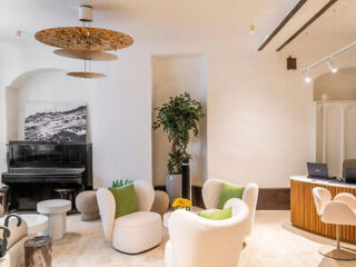 A modern lounge area with white armchairs, a piano, a potted plant, a desk with laptops, and ceiling light fixtures.