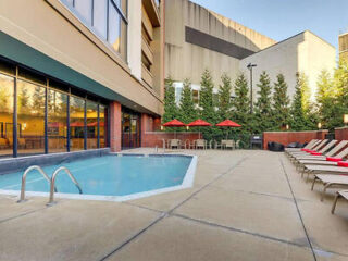 Outdoor pool area with lounge chairs, a small pool, red umbrellas over tables, and surrounding greenery. The setting is adjacent to a building and has clear skies.