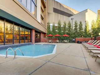 Outdoor pool area with lounge chairs, a small pool, red umbrellas over tables, and surrounding greenery. The setting is adjacent to a building and has clear skies.