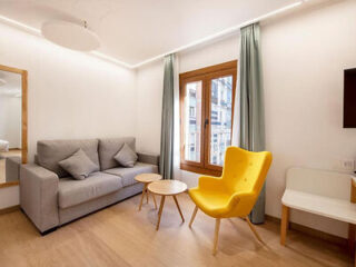 Modern living room with a grey sofa, yellow armchair, small wooden tables, a large window with curtains, and minimalistic decor.