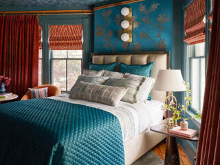 A bedroom with teal quilted bedding, a beige upholstered headboard, multiple pillows, red curtains, blue wallpaper with gold accents, a side table with a lamp, and two windows with striped shades.