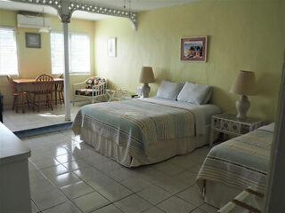 A well-lit room with pale yellow walls, two beds with striped bedspreads, two side tables, lamps, a wall painting, a small dining area with a table and four chairs, and a window with blinds.