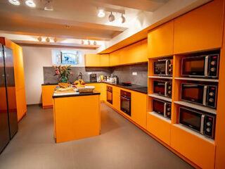 A modern kitchen with bright orange cabinets, multiple microwaves, and an island countertop, featuring various kitchen appliances and fresh fruit.