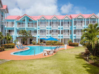 A bright and colorful resort-style building with a light blue exterior and red roofs, surrounding a central outdoor swimming pool with lounge chairs and umbrellas.