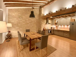 Modern kitchen and dining area with wooden beams, stone walls, and contemporary furniture. The space includes a wooden dining table, gray chairs, and a fully equipped kitchen with appliances.