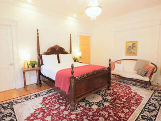 A bedroom with a large wooden four-poster bed, red and white bedding, a white loveseat with red pillows, and a patterned rug. There is a small nightstand with a lamp and plants on each side of the bed.