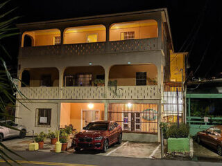 A two-story building with arched balconies is illuminated at night. A red car is parked in the driveway underneath. Several potted plants line the front of the building.