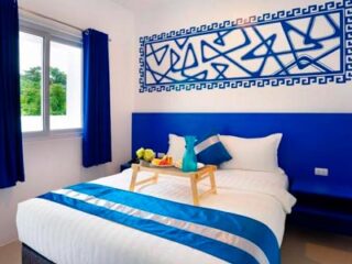 A neatly made bed in a hotel room with blue accents, a blue-patterned wall art, and a wooden tray with breakfast items.