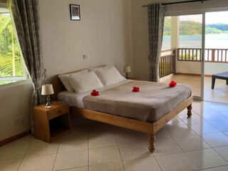 A simple bedroom with a wooden bed, decorated with red flower petals, near a large window and balcony overlooking greenery.