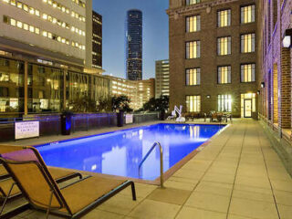 A rooftop pool at dusk, surrounded by lounge chairs, with tall buildings and a skyscraper in the background.