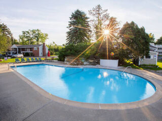 Outdoor swimming pool surrounded by lounge chairs with sunlight peeking through trees in the background.