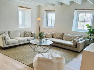 A living room with two beige sofas, a geometric glass coffee table, a tall floor lamp, a plant, and three windows. The room has white walls and a beige rug.