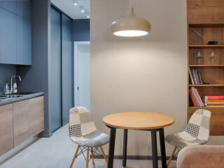 A small, modern dining area with two patterned chairs and a round wooden table, situated next to a minimalist kitchen and a bookshelf. A pendant light hangs above the table.