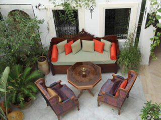 An outdoor courtyard featuring a wooden sofa with colorful cushions, surrounded by plants and traditional decor.