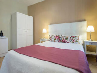 A neatly made bed with floral pillows, white bedding, and a pink throw blanket. There are two bedside tables with lamps, a white wardrobe, and a beige accent wall.