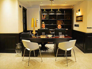 Hotel reception area with two staff members seated behind a dark wooden desk, under warm, elegant lighting.