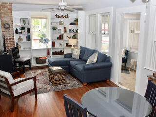 A cozy living room with a blue sectional sofa, wooden chairs, and a round glass dining table. Bookshelves, a ceiling fan, and large windows create a bright, inviting atmosphere.