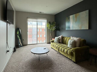 A modern living room with a green sofa, round coffee table, wall-mounted TV, large window with blinds, and abstract wall art. A potted plant and a full-length mirror are placed near the window.