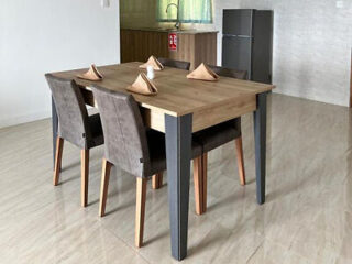 A minimalist dining area with a wooden table, four cushioned chairs, and folded napkins, with a small kitchenette in the background.