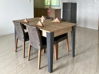 A minimalist dining area with a wooden table, four cushioned chairs, and folded napkins, with a small kitchenette in the background.