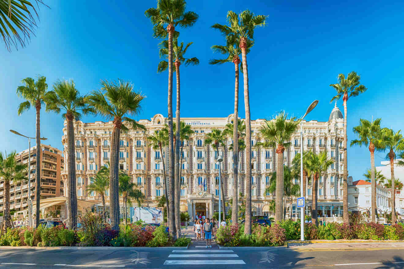 A grand, historic hotel with ornate architecture, lined with tall palm trees, and a lush garden in the foreground, under a clear blue sky.