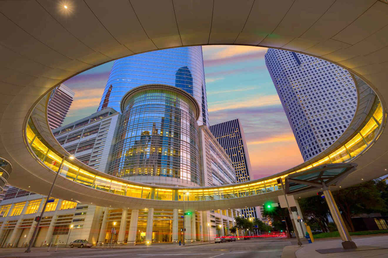 Circular overpass with glass-enclosed building and skyscrapers in background at sunset. Street below with cars and traffic lights.