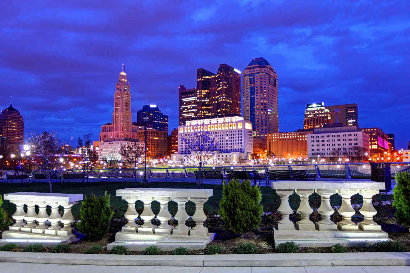 Cityscape at dusk featuring illuminated buildings against a deep blue sky, viewed from a nearby park with ornate stone fences and shrubbery in the foreground.