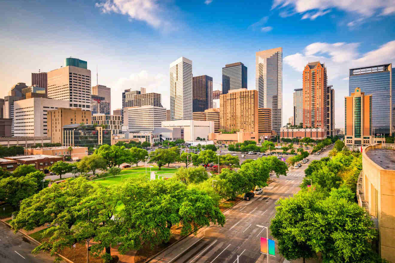 Downtown cityscape with modern skyscrapers, green trees, and a street with some vehicles under a clear blue sky.