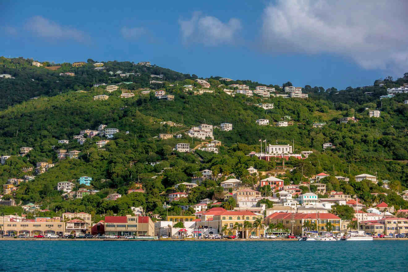 Hillside view of a coastal town with numerous buildings among green vegetation, clear blue sky, and calm sea in the foreground.