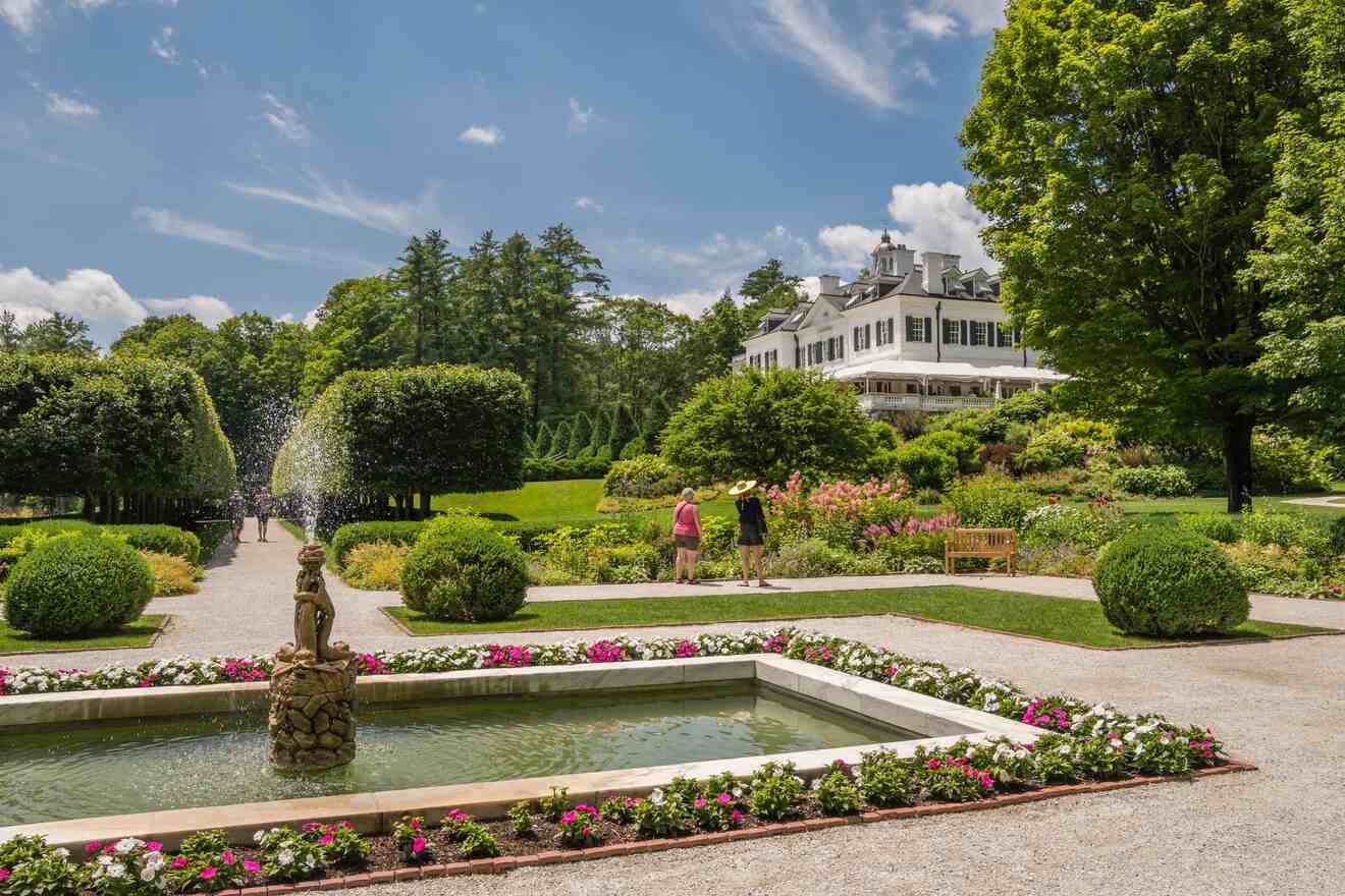 A landscaped garden features a rectangular fountain with a statue, colorful flower beds, and two people walking towards a large white mansion in the background under a blue sky.