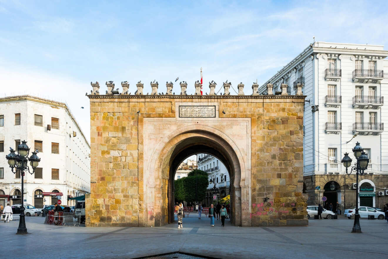 The historic Bab el Bhar gate in Tunis, with pedestrians walking underneath its stone archway and surrounded by classic buildings.