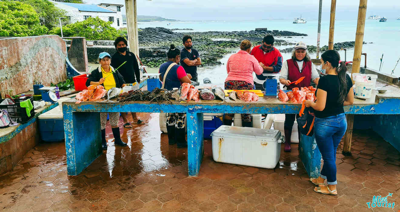 People at an outdoor market stall process and display fish near a rocky shoreline.