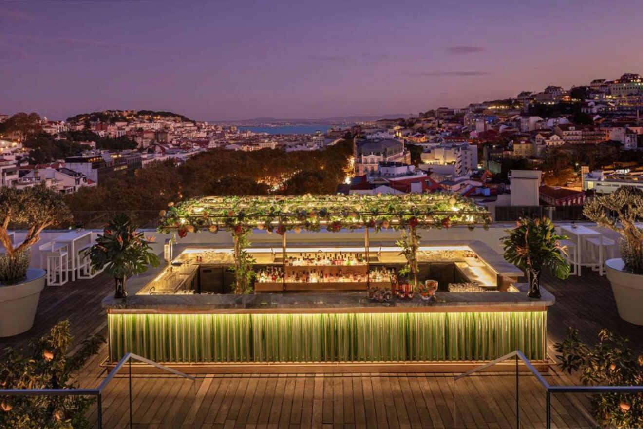 An illuminated rooftop bar with greenery overlooks a cityscape at dusk, featuring seating areas and a distant view of rolling hills and a body of water.