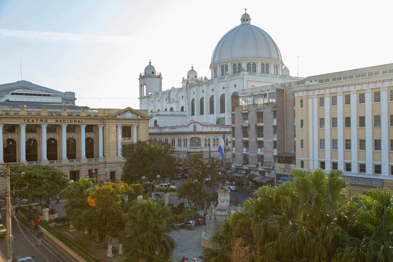 A city square with a park surrounded by buildings, including the Teatro Nacional and a white domed structure. Trees and a statue are visible in the foreground.