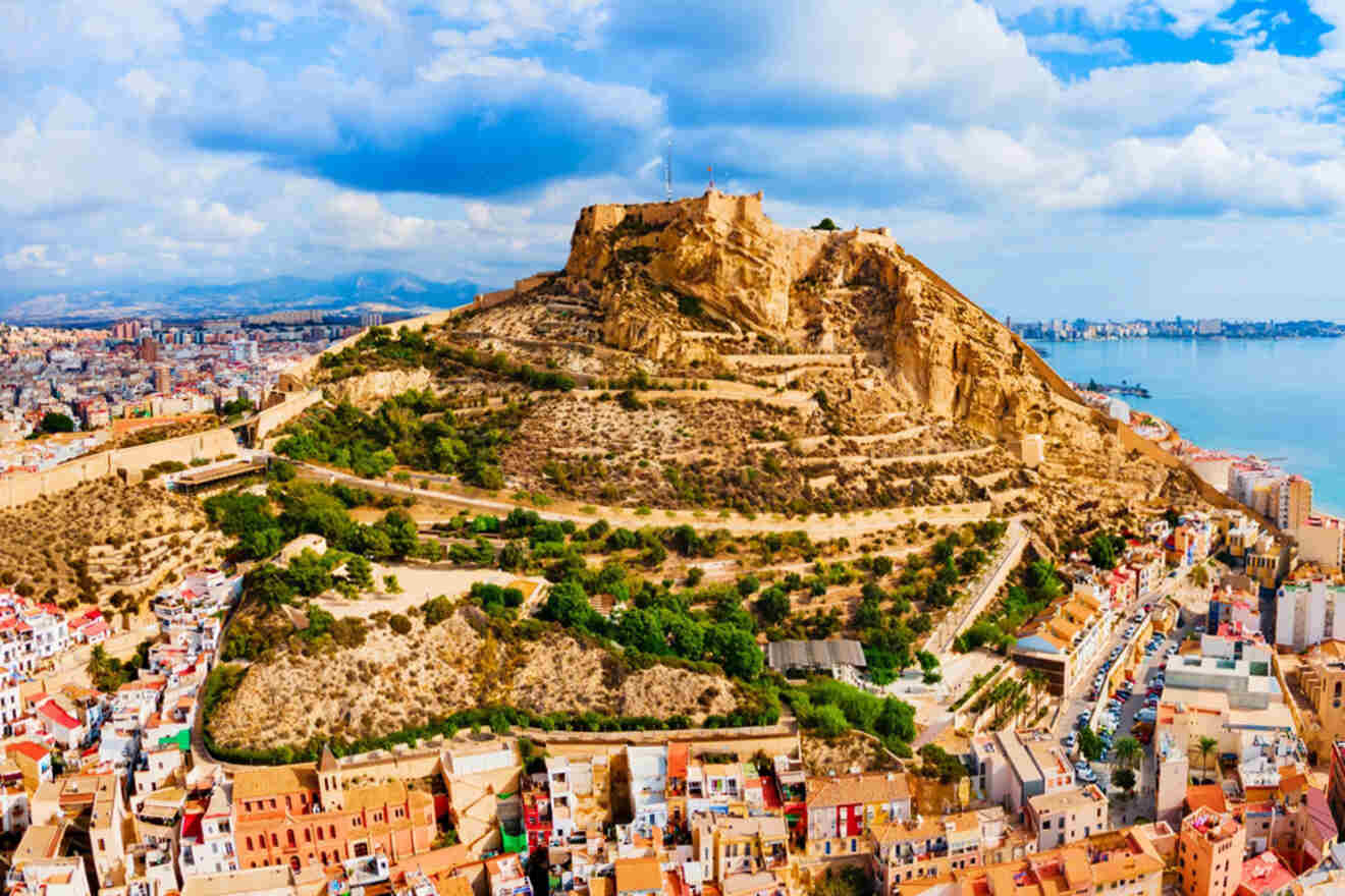 Aerial view of a hilltop fortress surrounded by an urban landscape, with a mix of modern and traditional buildings, and coastline visible in the background under a partially cloudy sky.