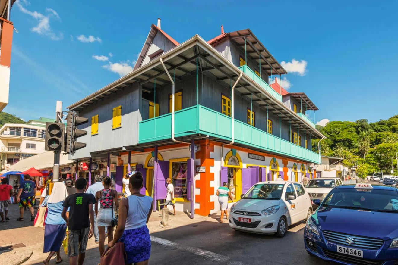 Street scene showing pedestrians walking past a colorful two-story building with shops on the ground floor and a car parked in front. A clear blue sky is in the background.